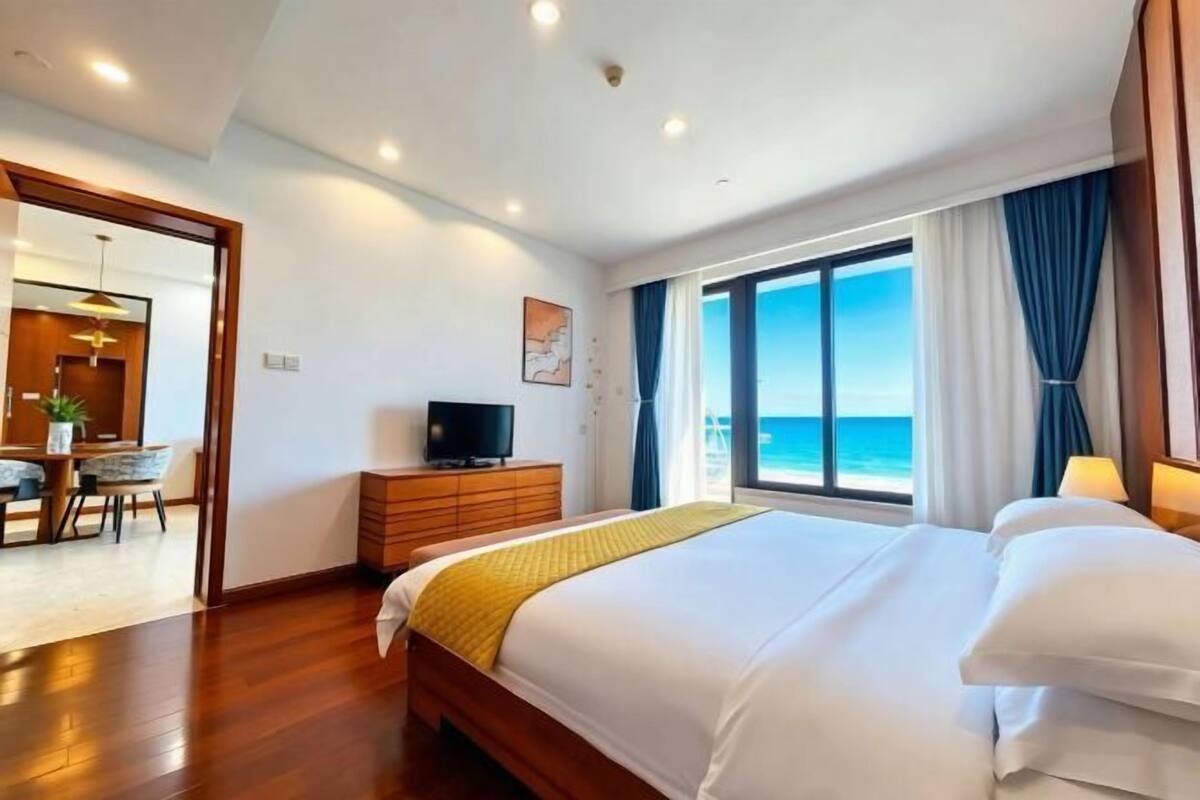 A hotel room with a large bed, a TV on a wooden dresser, a desk with a chair, and a view of the ocean through large windows.