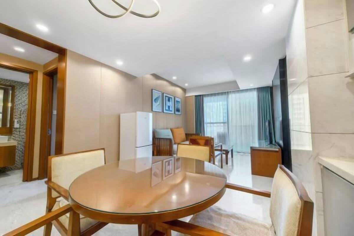 A modern hotel room with a round glass table, wooden chairs, a sofa, a TV, and a large window with curtains.