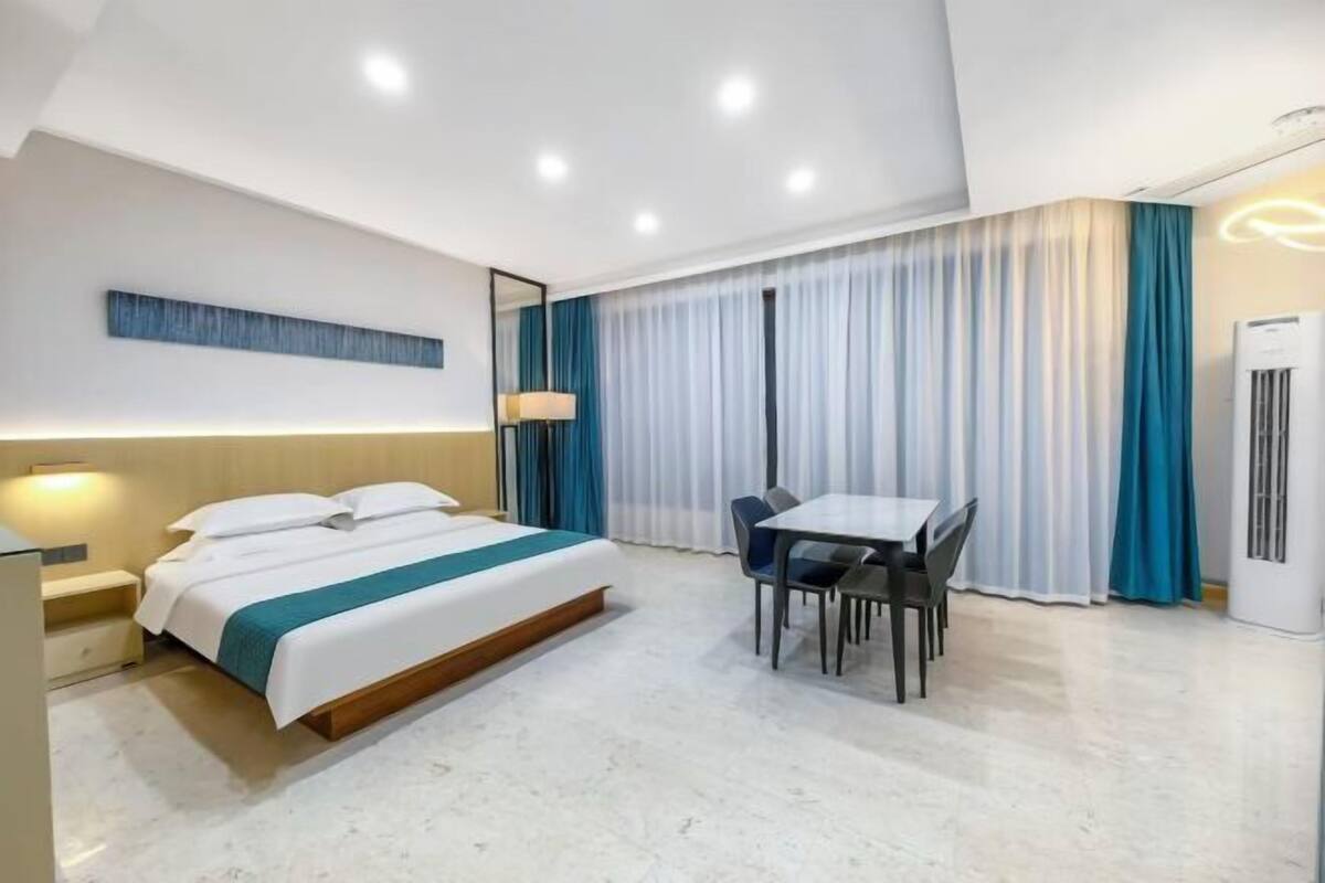A modern hotel room with a bed, bedside table, lamp, dining table with chairs, and a large window with curtains.