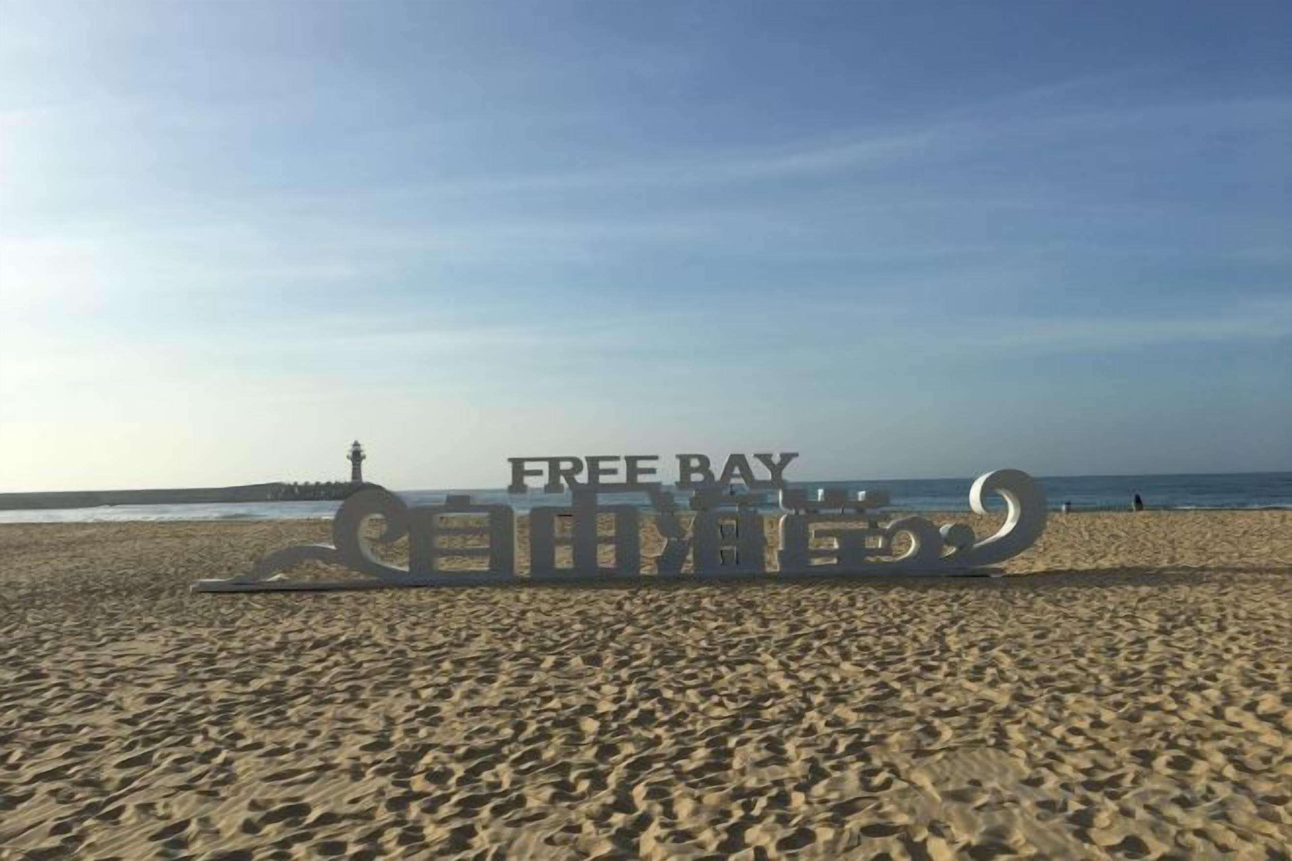 A beach with a large metal sign reading 'FREE BAY' and a lighthouse in the background.
