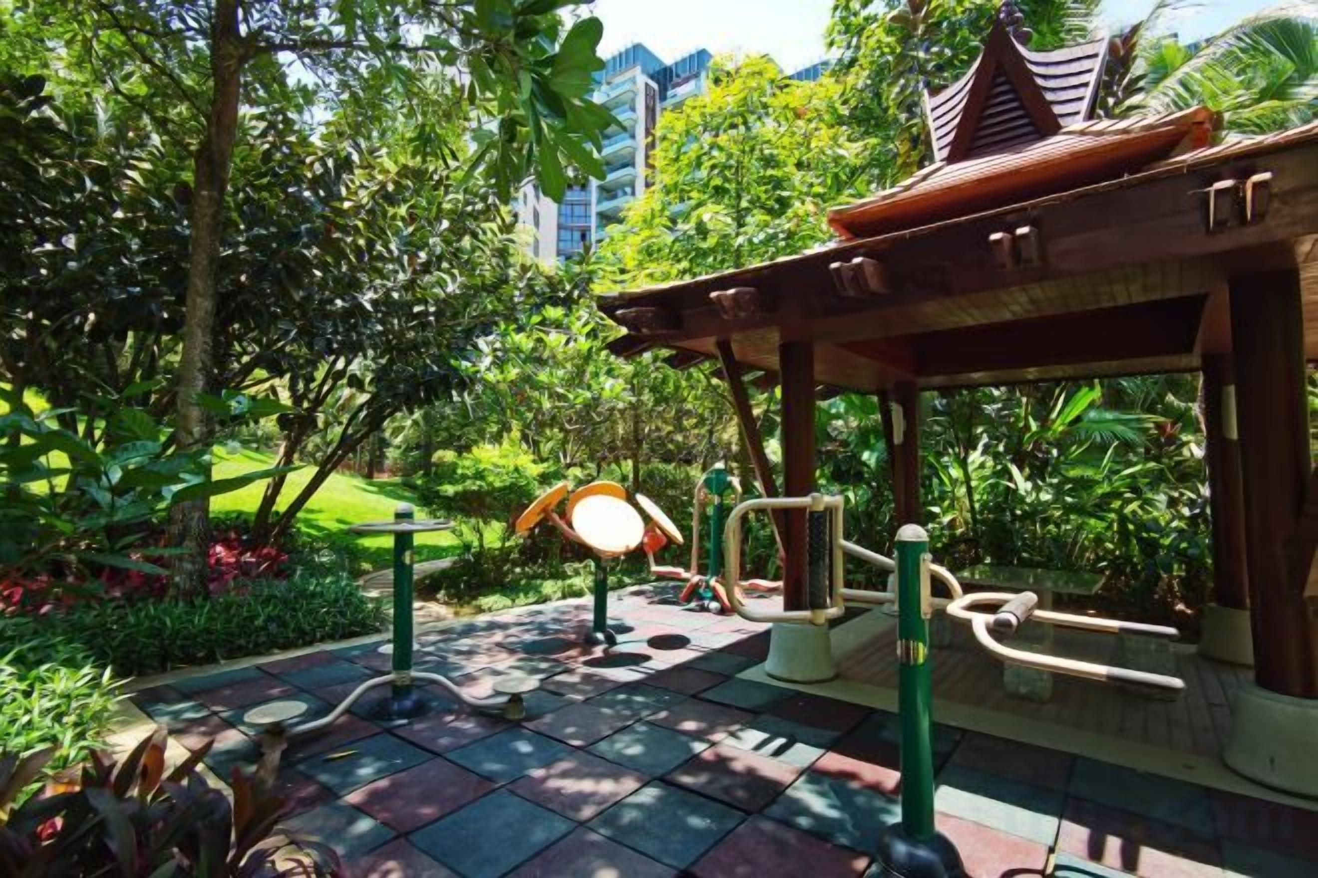 A covered outdoor area with exercise equipment, surrounded by lush greenery and a tiled floor.