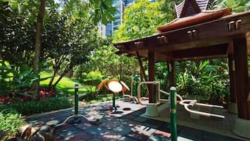 A covered outdoor area with exercise equipment, surrounded by lush greenery and a tiled floor.