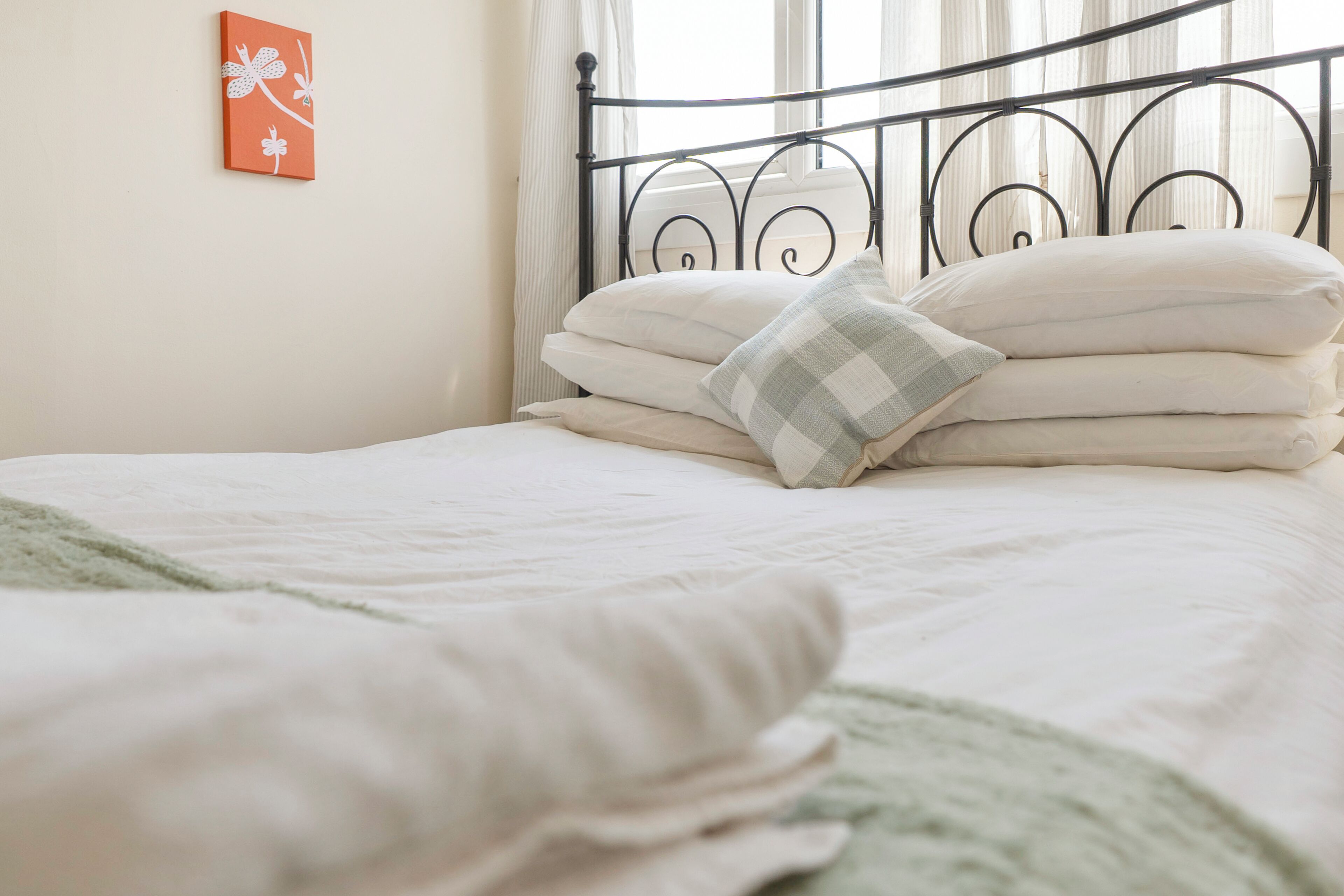 A neatly made bed with white bedding and a checkered pillow, a metal headboard, and a wall with a framed picture.