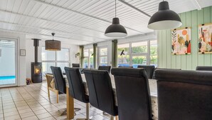 Un comedor moderno con una mesa larga y sillas negras, un estufa de leña y ventanas amplias con vistas al follaje.