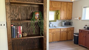 A wooden bookshelf with books and decorative items, a kitchen with wooden cabinets and a microwave, and a ceiling light fixture. - Cozy acorn cottage w/ lake view (West Branch)