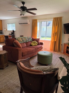 A living room with a red sofa, a green ottoman, a wicker chair, a ceiling fan, and a television.