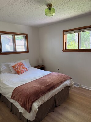 A bedroom with a bed, pillows, a nightstand, a lamp, and a window with blinds.