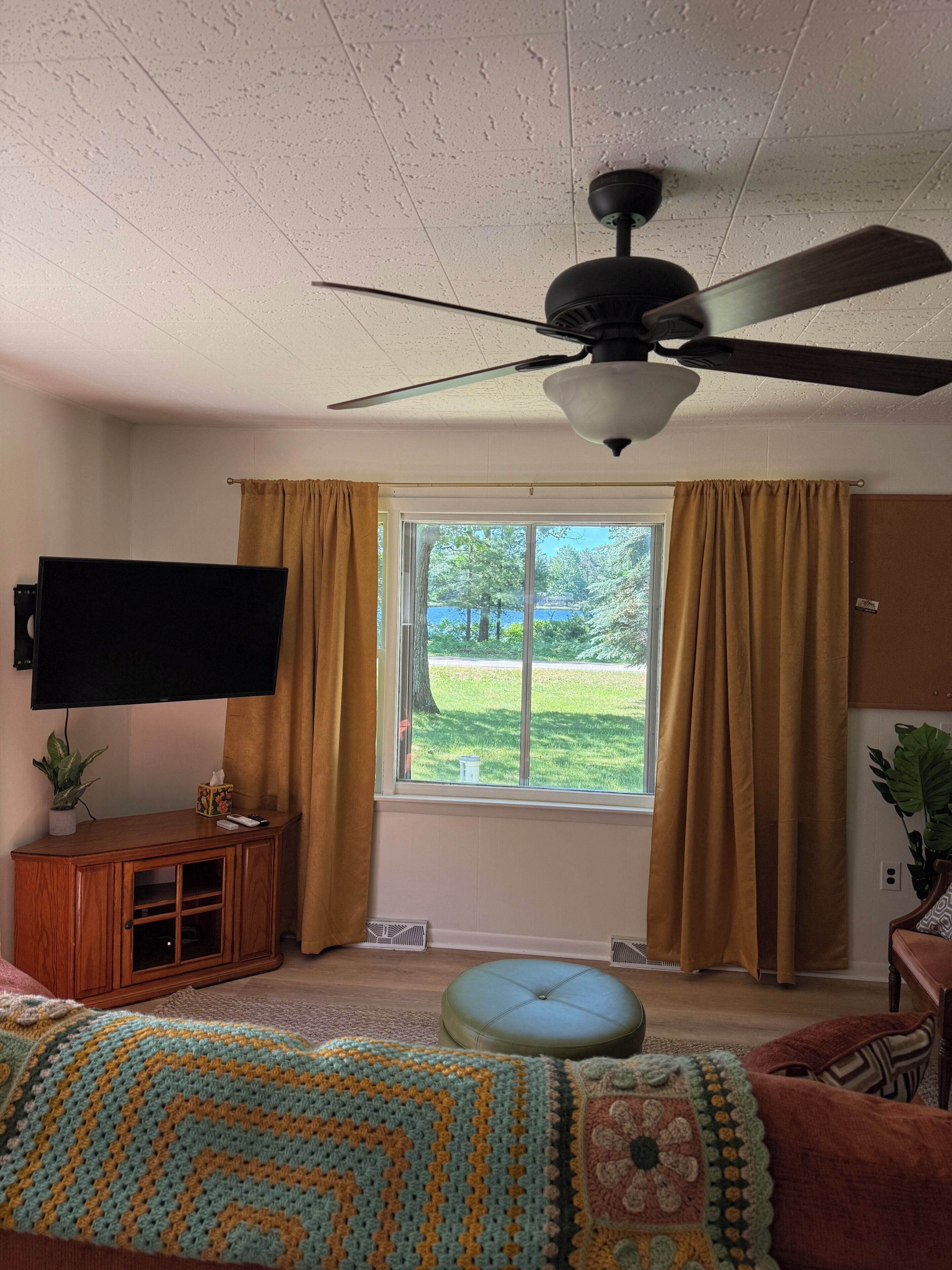 A room with a ceiling fan, a television, a window with curtains, a wooden cabinet, and a large window offering a view of a lawn.