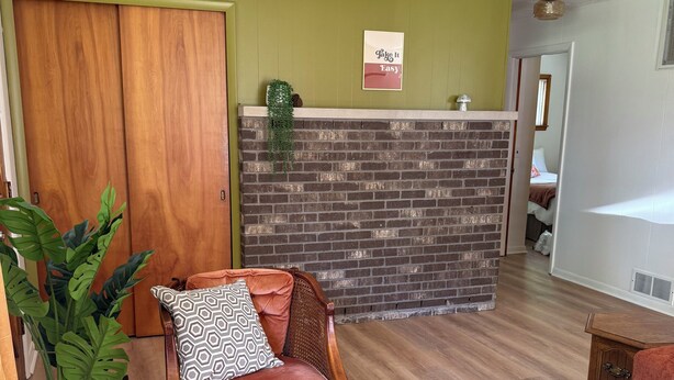 A living room with a wooden door, a brick accent wall, a cushioned chair, a green ottoman, and a woven rug. - Cozy acorn cottage w/ lake view (West Branch)