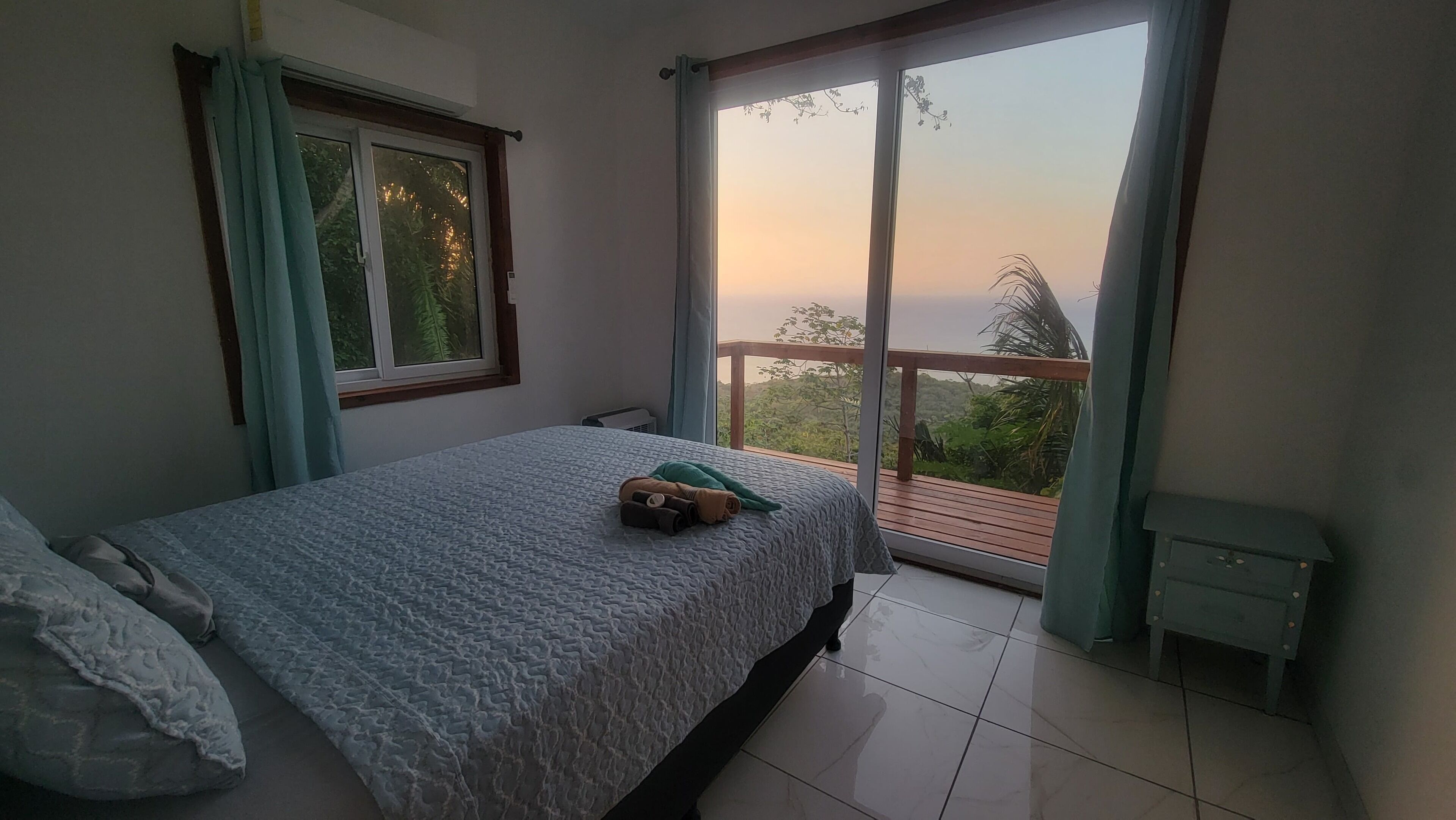 A bedroom with a bed, a window, a balcony, and a view of greenery.