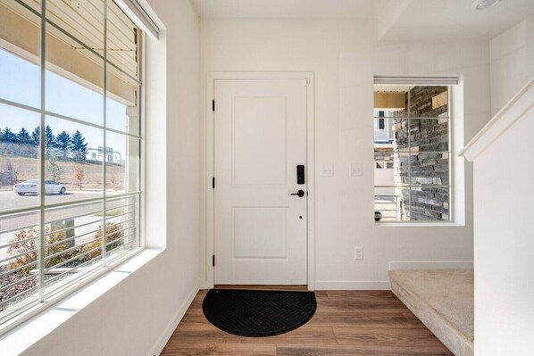 Un couloir avec une porte blanche, une petite fenêtre donnant sur un immeuble et un paillasson noir.