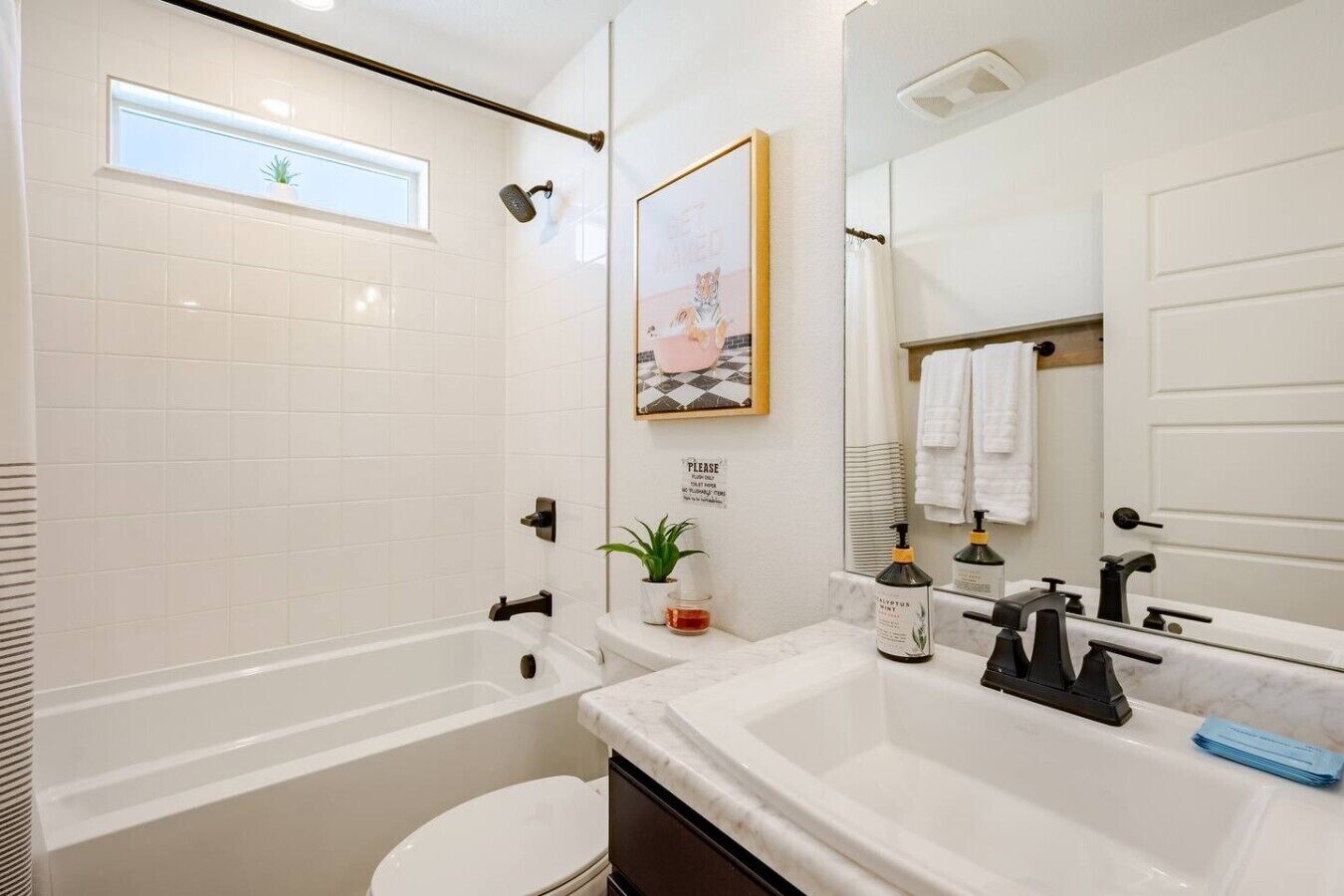 Een badkamer met een bad, toilet, wastafel en douche. Er is een raam, een pot met een plant en een lijst met een foto aan de muur.