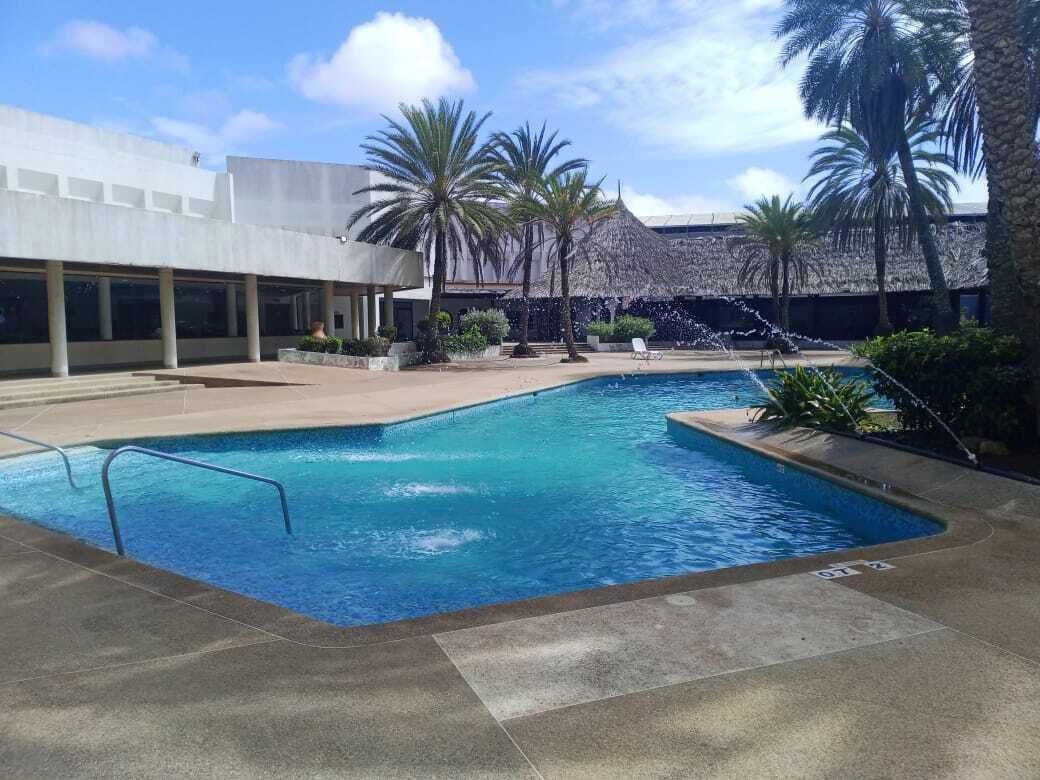 A swimming pool with a water fountain, palm trees, and a modern building in the background.