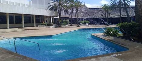 A swimming pool with a water fountain, palm trees, and a modern building in the background.