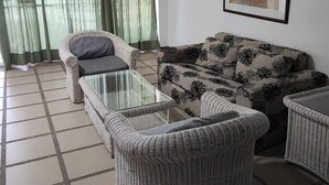Een woonkamer met een bank, twee fauteuils, een glazen salontafel en een groot raam met groene gordijnen.