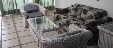 Un salon comprenant un canapé, deux fauteuils, une table basse en verre et une grande fenêtre ornée de rideaux verts.