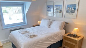A neatly made bed with white linens and towels, a bedside table with a lamp, and a colorful rug on a wooden floor.