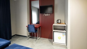 Ein Hotelzimmer mit einem Einzelbett, einem Schreibtisch mit Stuhl, einem an der Wand montierten Fernseher und einem kleinen Schrank mit Wasserkocher.