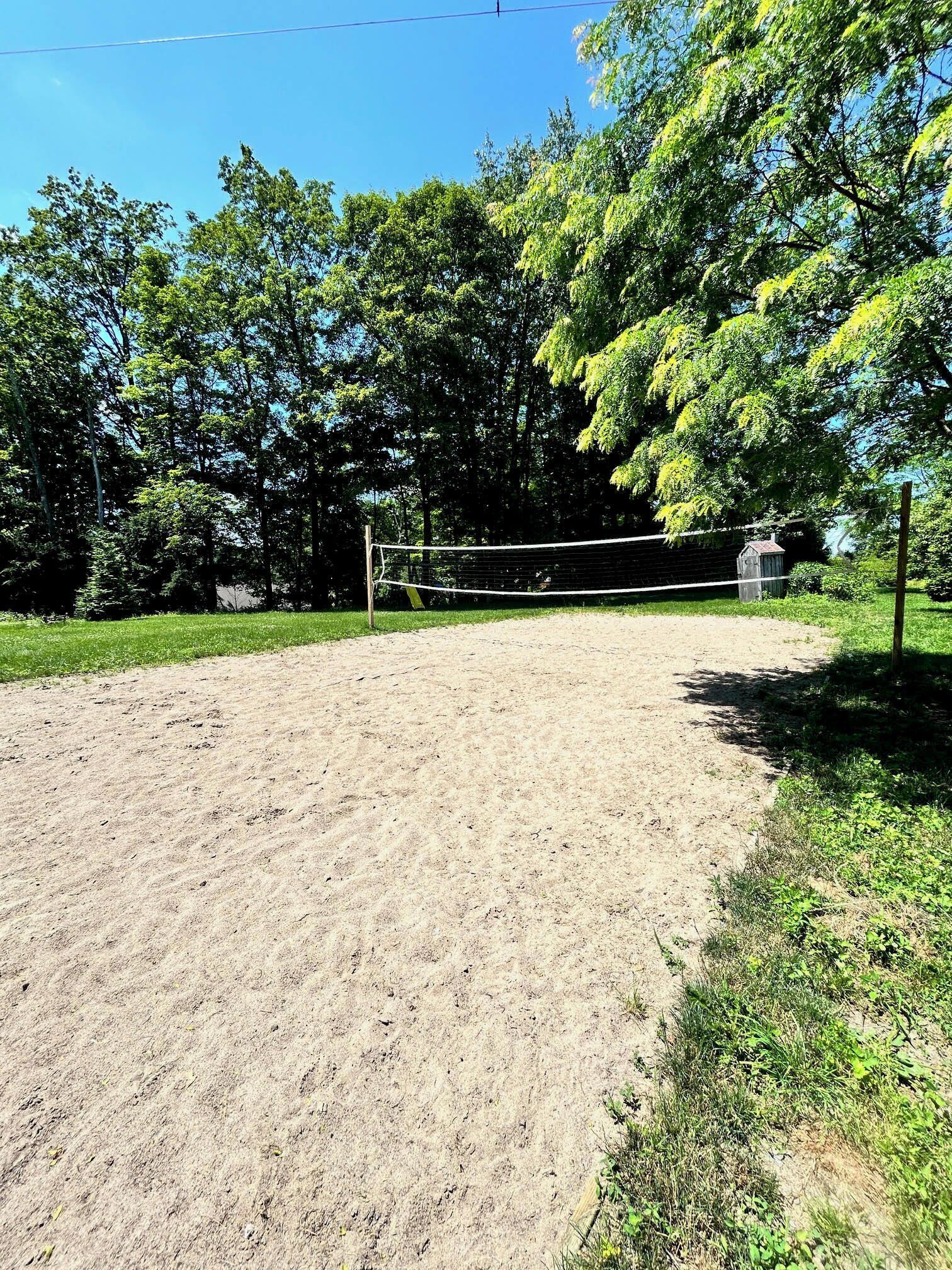En sandvollybollplan med ett nät, omgiven av träd och en träkonstruktion i bakgrunden.