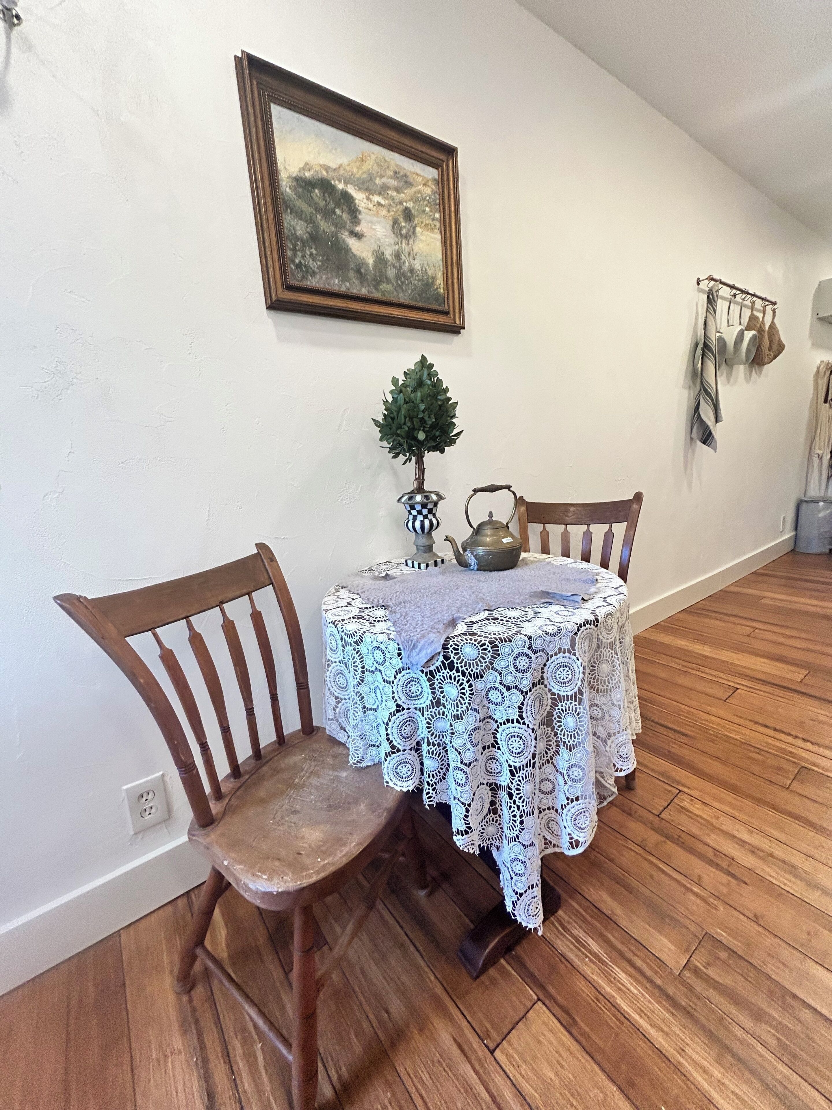 Un coin repas avec une table recouverte d’une nappe en dentelle, deux chaises en bois, une petite plante en pot et un tableau accroché au mur.