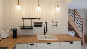 Cocina con encimera de madera, lavamanos blanco y tres lámparas colgantes. Se observan dos cestas colgantes negras y un grifo negro. En el fondo, hay una tostadora y una lavadora.