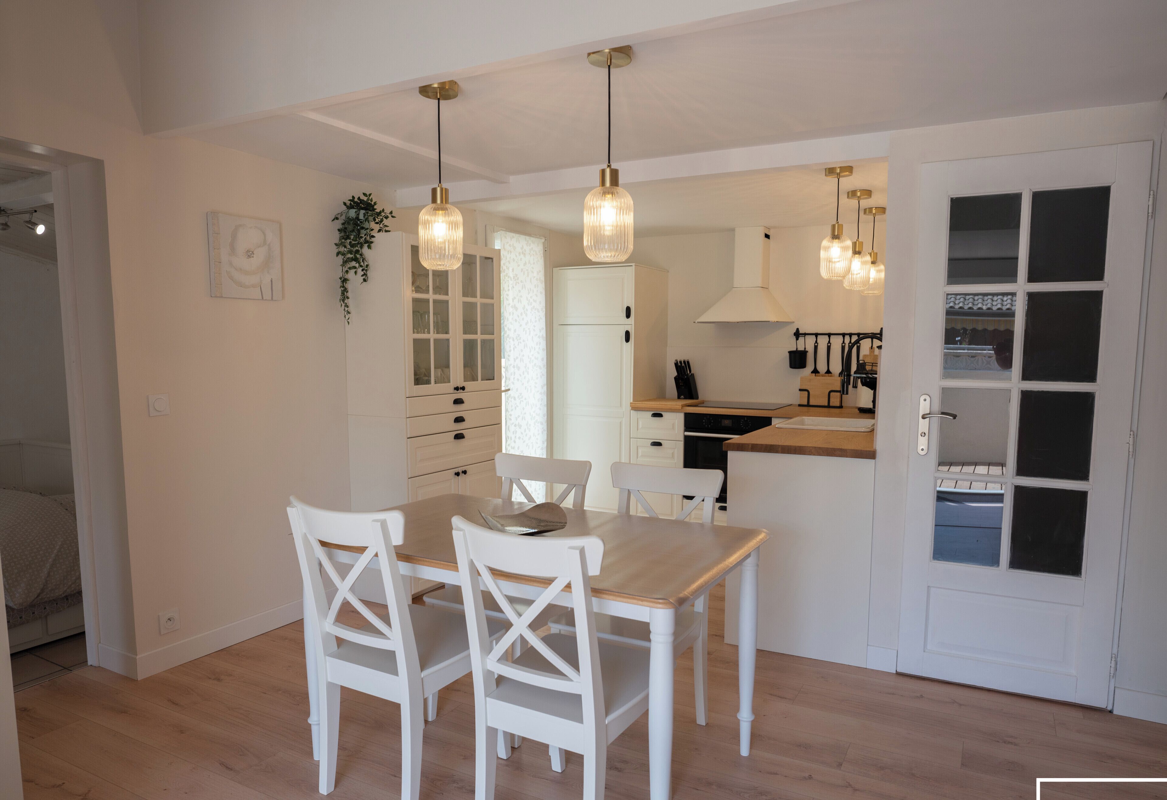 Una cocina moderna con armarios blancos, una mesa de comedor con sillas, lámparas colgantes y vistas al dormitorio contiguo.