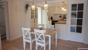 Una cocina moderna con armarios blancos, una mesa de comedor con sillas, lámparas colgantes y vistas al dormitorio contiguo.