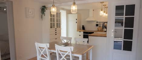 Una cocina moderna con armarios blancos, una mesa de comedor con sillas, lámparas colgantes y vistas al dormitorio contiguo.