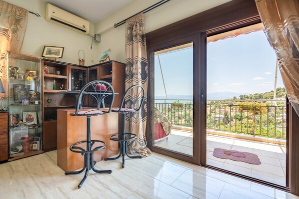 A room with a wooden desk, two black chairs, a glass cabinet with various items, a large sliding glass door to a balcony with a view of greenery and mountains. - Peaceful Hilltop Apartment With Balcony Sea View (Chalcis)