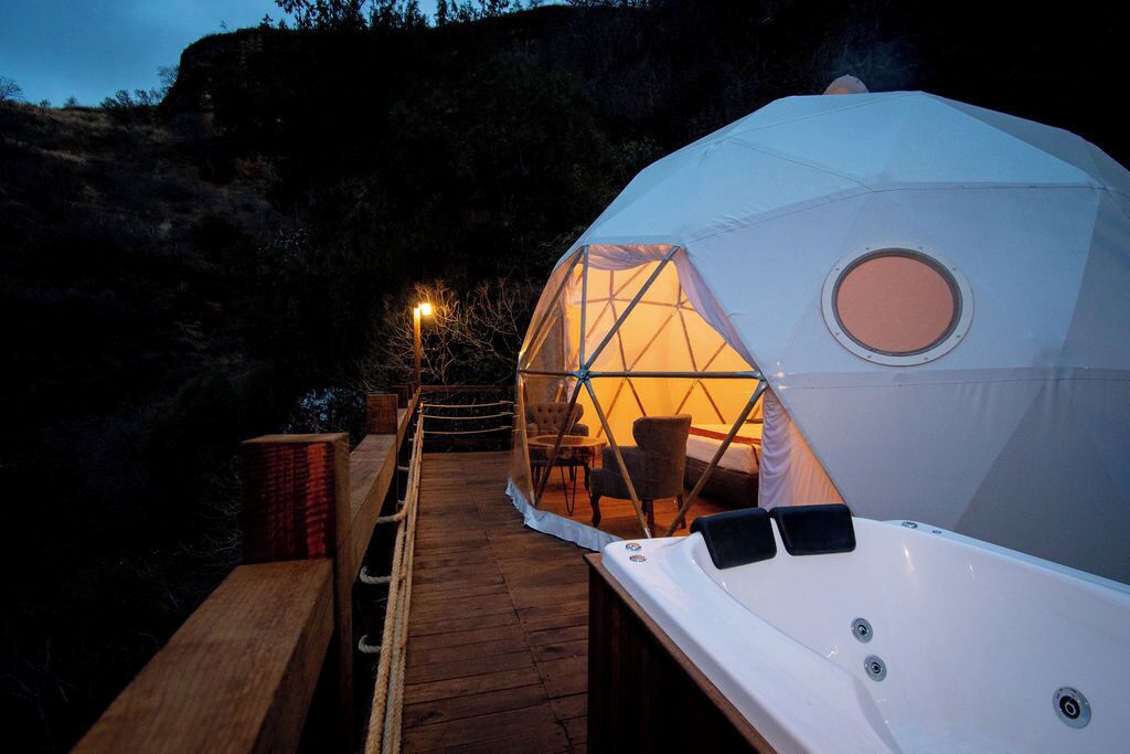 Estructura de domo geodésico con una terraza de madera que conduce a él, un ventanal circular y una silla en su interior.