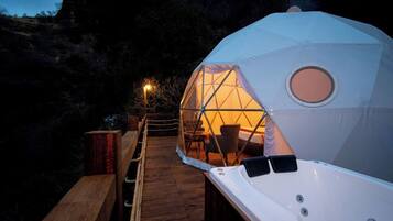 Estructura de domo geodésico con una terraza de madera que conduce a él, un ventanal circular y una silla en su interior.