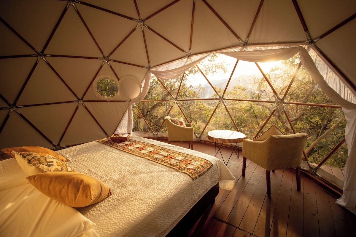 Un dormitorio tipo tienda de campaña, acogedor y triangular, con cama, almohadas, una mesita y sillas.