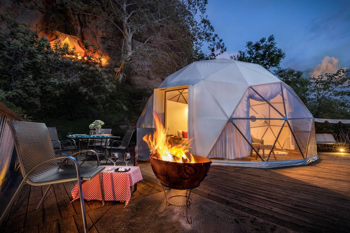 Una tienda en forma de domo geodésico con un brasero en el centro, rodeada por una terraza de madera y mobiliario exterior.
