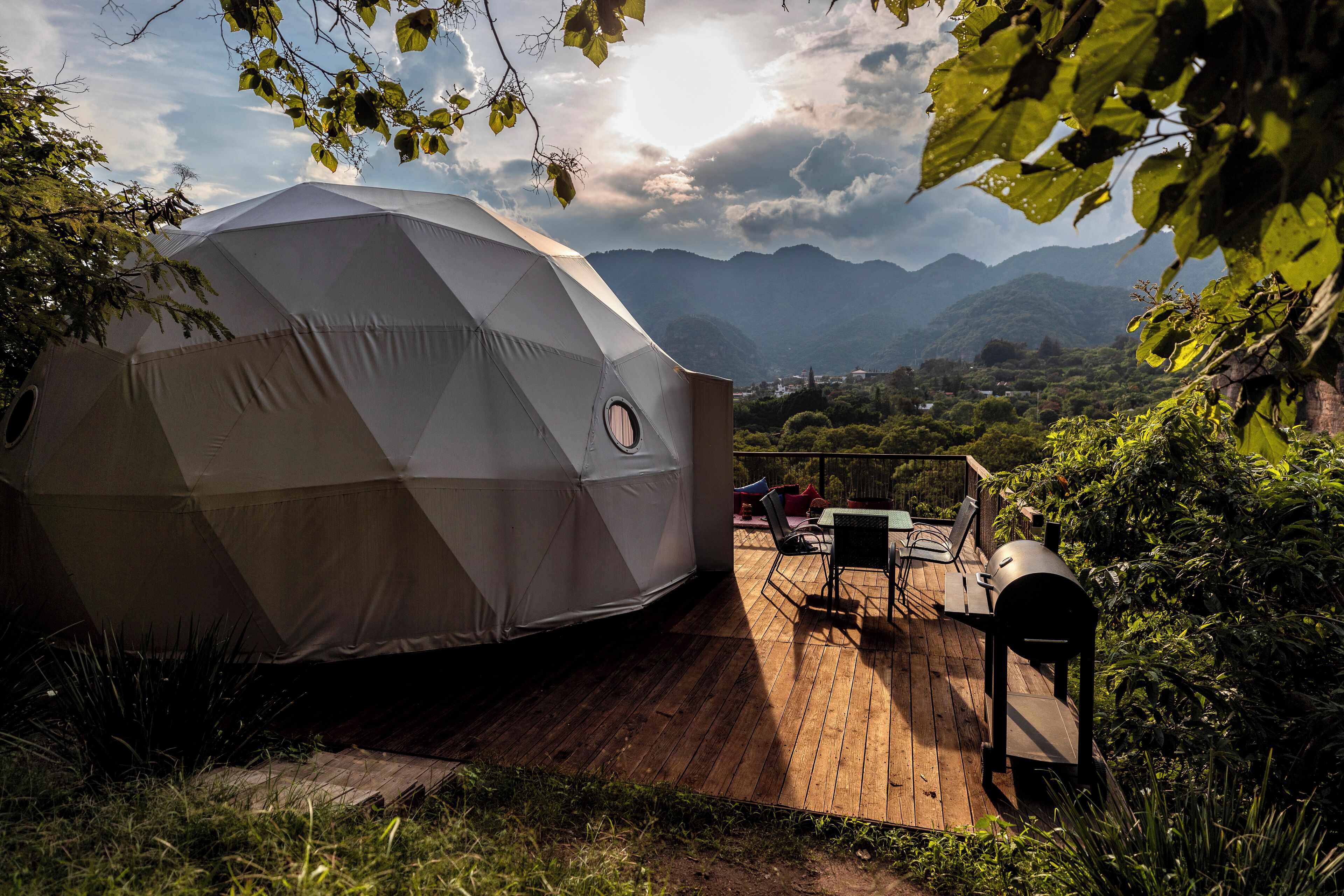 Uma tenda em forma de cúpula geodésica com deck de madeira e churrasqueira, rodeada por vegetação exuberante e montanhas.