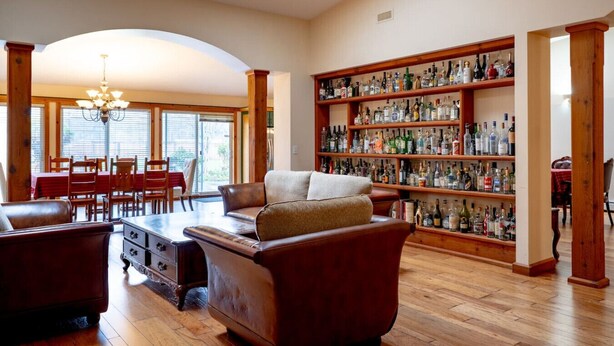 A spacious living room with a large leather sofa, a wooden coffee table, and a dining area with a red tablecloth. A large bookshelf filled with various bottles and glasses is visible in the background. - Fort Wine Manor: Entire Private Estate Home (Langley Township)