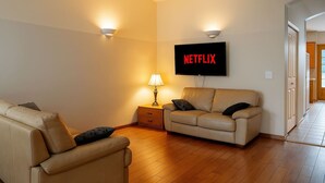 A living room with a beige sofa, a smaller armchair, a lamp, and a wall-mounted TV displaying "NETFLIX". - Fort Wine Manor: Entire Private Estate Home (Langley Township)
