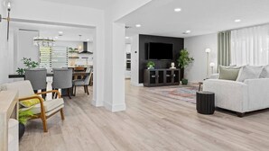 A modern living room with a sofa, a dining area with chairs, a kitchen with a TV, and a large window with curtains.