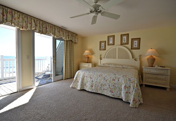 A bedroom with a bed, two bedside tables with lamps, a ceiling fan, a balcony with a view, and framed pictures on the wall.