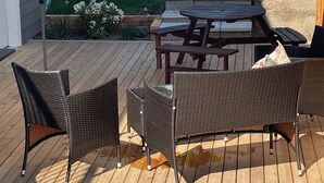 Eine Terrasse im Freien mit Holztisch und Korbstühlen, einem Gartenschlauch und einem Gartenbereich mit Rasen und Pflanzen.