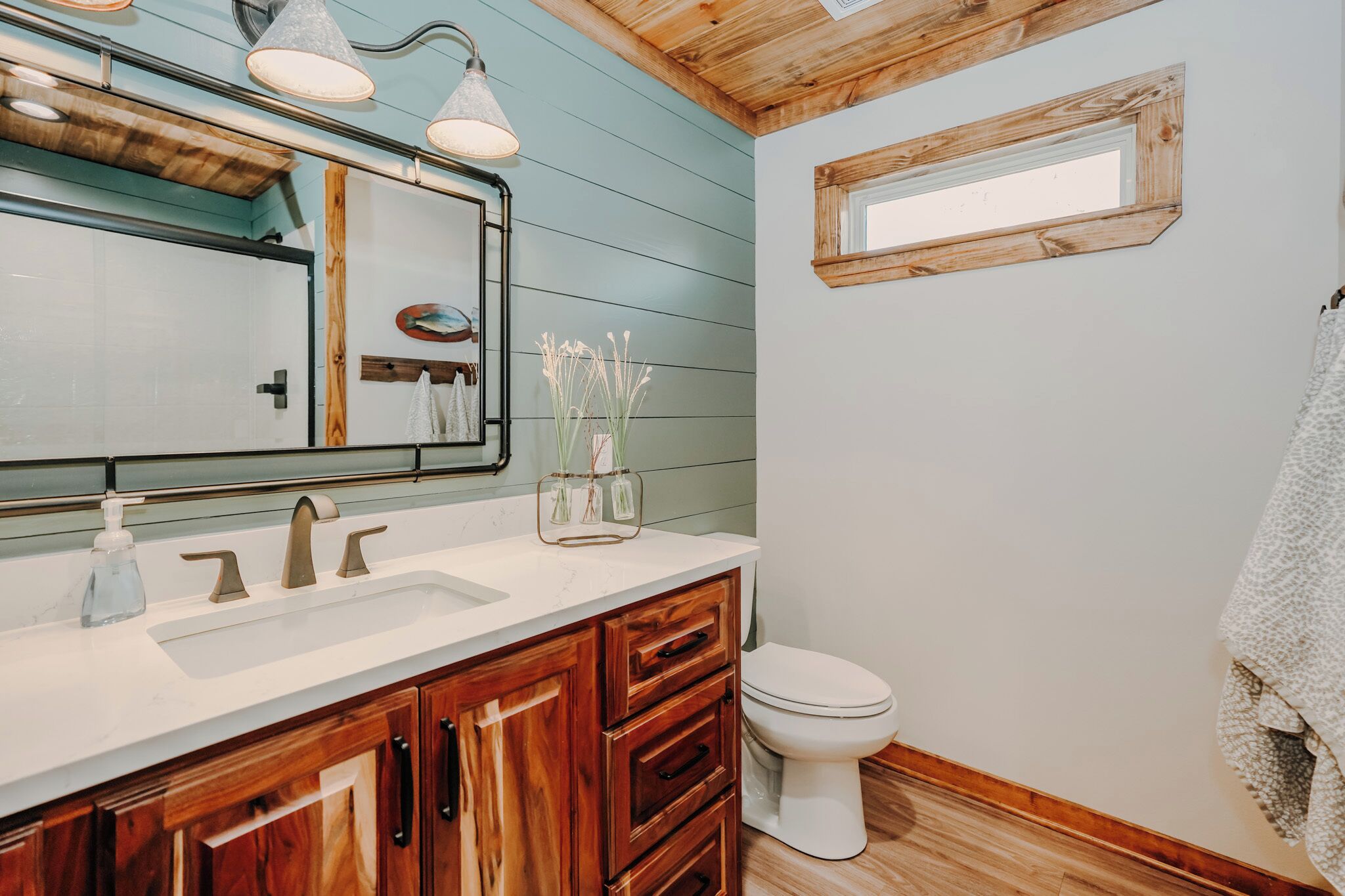 A bathroom with wooden cabinets, a white sink, and a toilet. There is a mirror with a wooden frame and a window above the toilet.