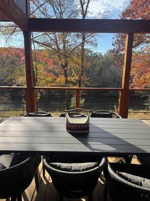 Un espace extérieur aménagé pour les repas, avec une table et des chaises, une rambarde en bois et une vue sur des arbres aux couleurs automnales.