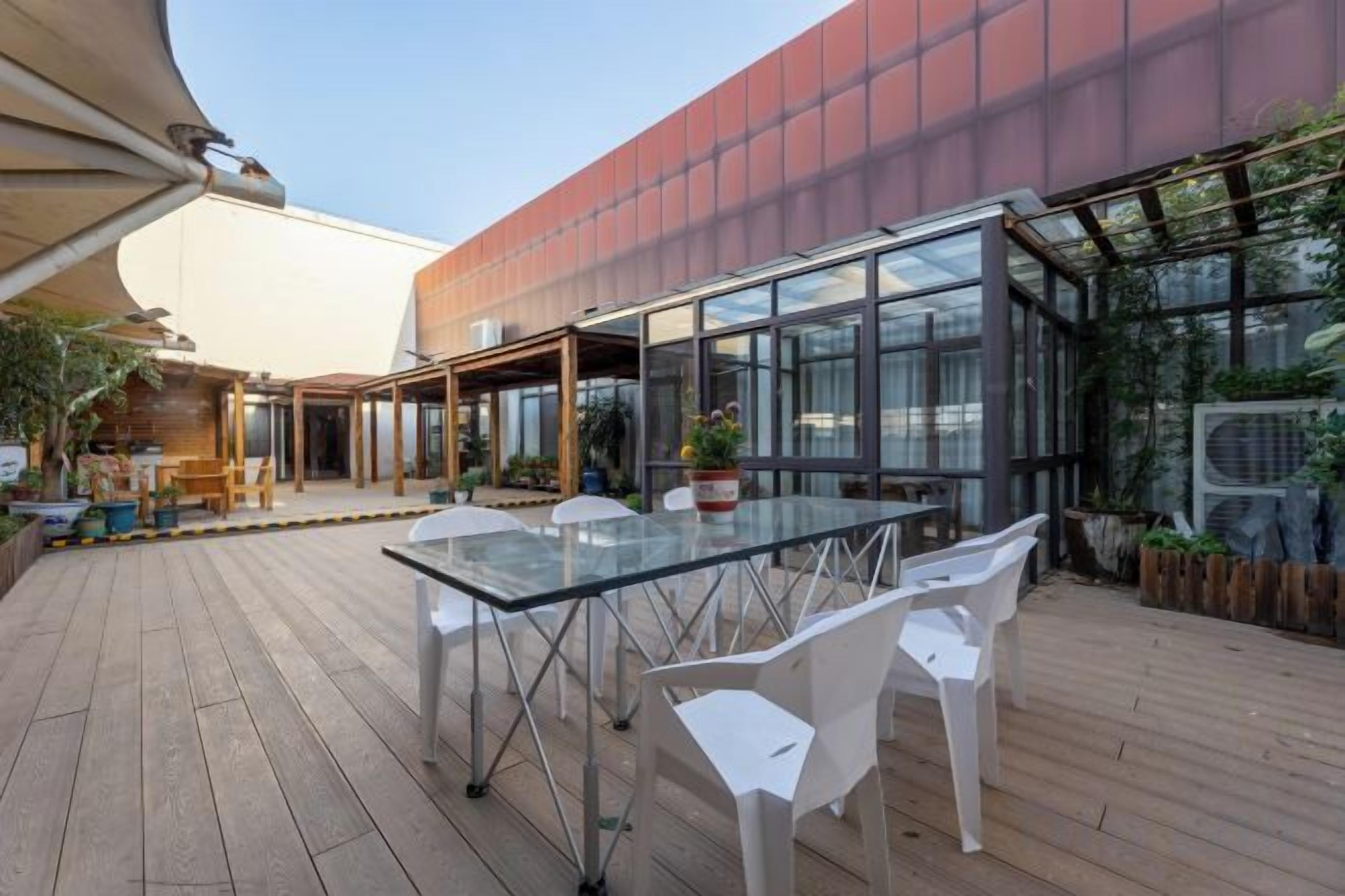 Un patio al aire libre con una mesa y sillas de vidrio, una cubierta de madera y un edificio moderno con grandes ventanales.