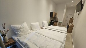 A hotel room with two beds, a small desk, a TV mounted on the wall, and a kitchenette area.