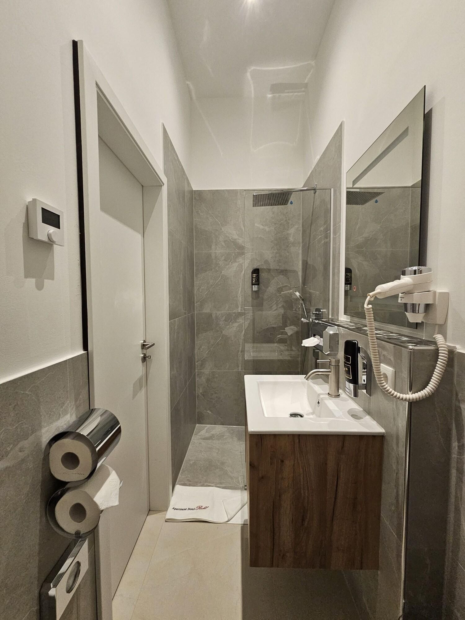Ein modernes Badezimmer mit Dusche, Waschbecken und Toilette. Toilettenpapierrollen und ein Handtuchhalter sind sichtbar.