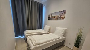 A small, modern bedroom with two single beds, a large window with curtains, a cityscape painting on the wall, and a potted plant.
