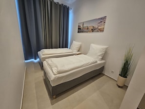 Une petite chambre à coucher moderne, avec deux lits simples, une grande fenêtre avec des rideaux, un tableau représentant un paysage urbain accroché au mur et une plante en pot.