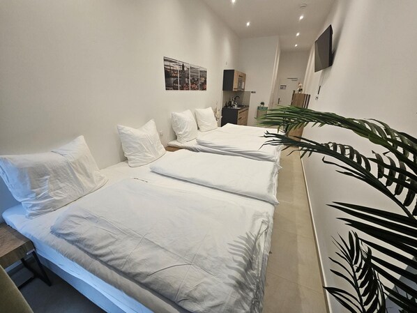 Une chambre d’hôtel avec trois lits, une grande plante en pot et une œuvre d’art représentant un paysage urbain accrochée au mur.