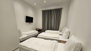 A hotel room with two single beds, a desk, a chair, a flat-screen TV mounted on the wall, and a window with grey curtains.