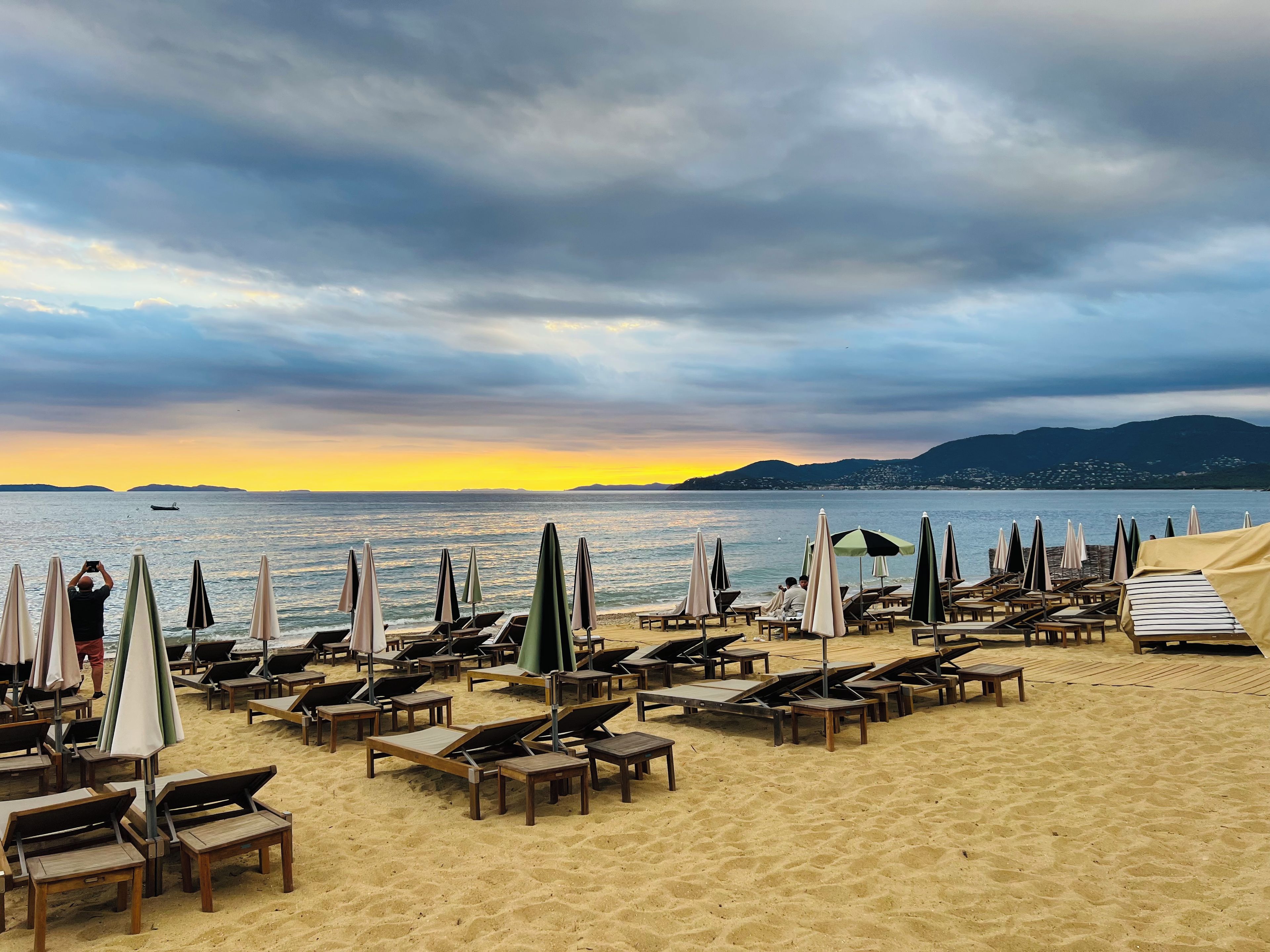 A beach with rows of sun loungers and umbrellas, a clear view of the sea, and distant mountains.