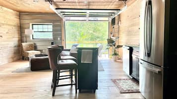A wooden interior with a kitchen area, a dining table with chairs, and a view through a large window.